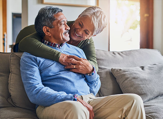 An elderly couple sits on a couch in a cozy living room.