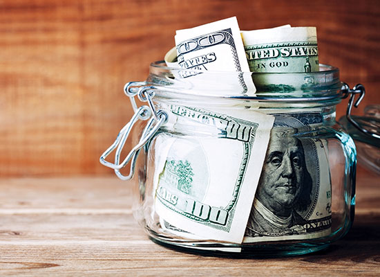 A glass jar filled with US dollar bills, mostly hundreds, sits as a testament to retirement dreams on a wooden surface