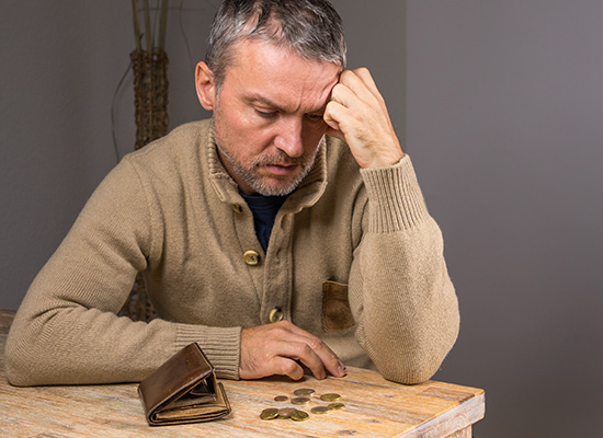 A middle-aged man sits at a table with his head in his hand, looking stressed as he examines a nearly empty wallet and some coins, indicating financial difficulties.