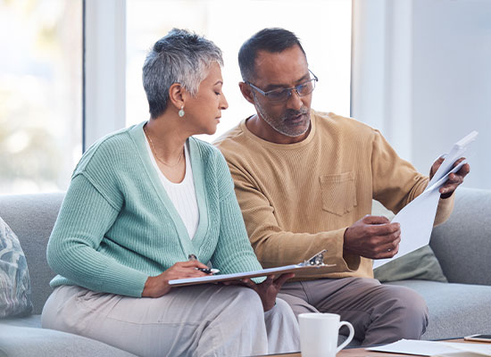 An older couple sits on a sofa. The woman, holding a clipboard and pen, looks at the man as he examines papers related to Social Security benefits