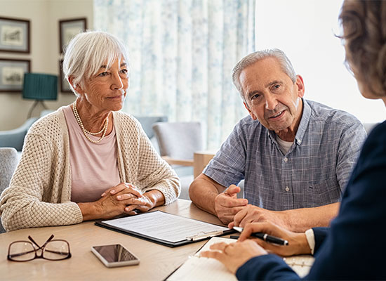 An elderly couple sits at a table with a financial advisor, discussing retirement planning. The woman, with short white hair, listens attentively while the man gestures as he speaks