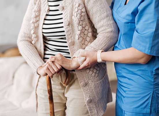 A person wearing a striped shirt and knitted cardigan uses a cane while being supported by someone in a blue uniform