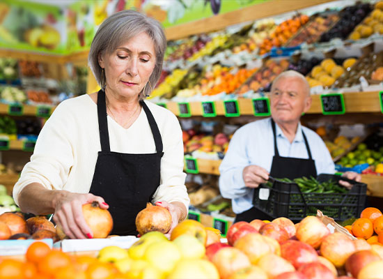 A woman with gray hair, wearing a white shirt and black apron, sorts fruit on a market stall. Behind her, an older man with white hair and a black apron holds a crate of vegetables, looking at her.