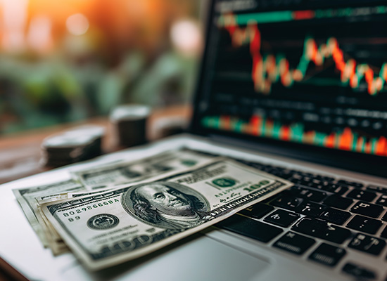 A laptop displaying financial charts with a few hundred-dollar bills on the keyboard, set against a blurred background, representing the integration of modern technology in financial monitoring and analysis.