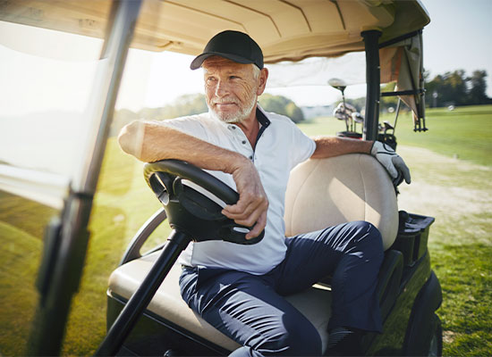 An older man with a white beard and wearing a black cap sits in a golf cart, leaning on the steering wheel. Dressed in a white polo shirt, dark pants, and a golf glove on one hand, he epitomizes the baby boomers enjoying their peak 65 retirement surge on a sunny golf course.
