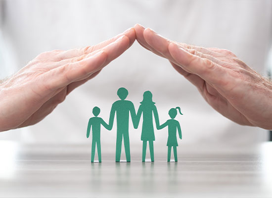 Two hands form a protective roof over a cutout family of four, symbolizing the concept of life insurance and family protection. The green figures, representing two adults and two children, rest on a light-colored surface, conveying security and peace of mind.