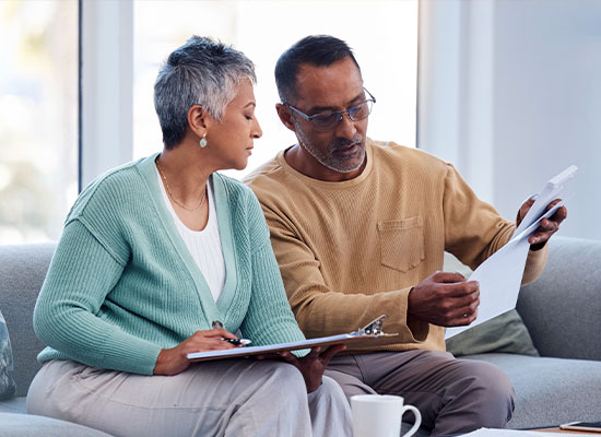 A senior couple sits on a couch at home. The woman, wearing a green cardigan, holds a clipboard and looks intently at the man, who is showing her a document about estate planning.