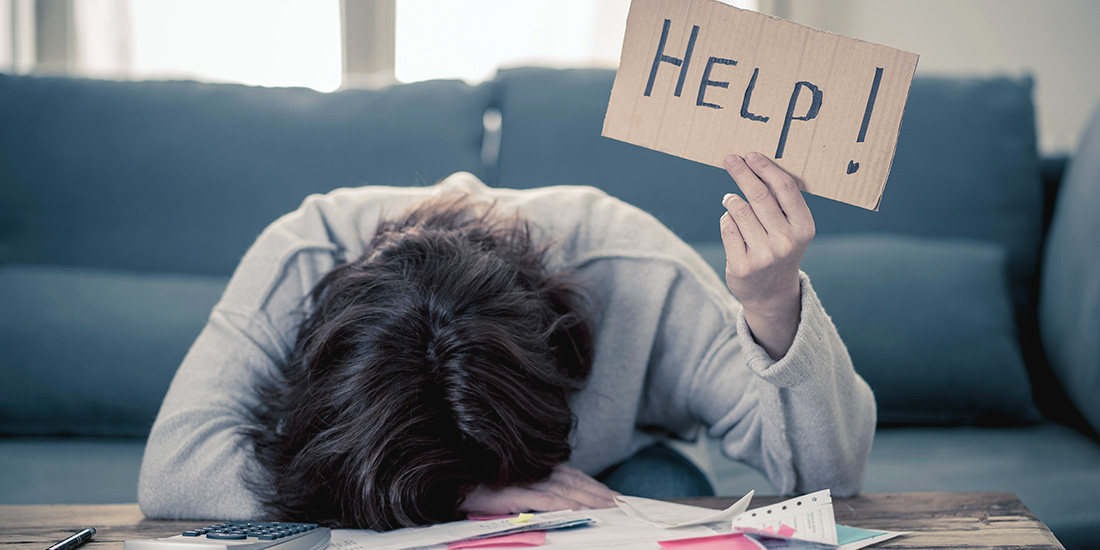 Woman with her down on a desk full of paperwork holding up a HELP sign