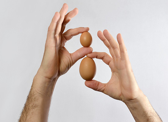 Two hands manipulating an egg in a balancing act, with one hand above the other against a plain light grey background.