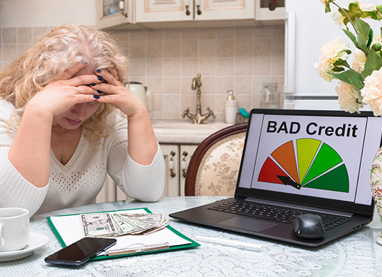 A person holds their head in frustration while sitting at a table with a laptop showing 'BAD Credit' and a credit score meter, suggesting struggles with poor credit rating.