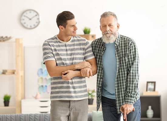 A young man supports his aging father, who uses a cane, as they walk through a bright living room, representing the caregiver role and assistance with activities of daily living.
