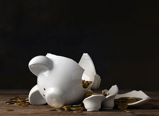 A broken white piggy bank with coins scattered around it on a wooden surface, against a dark background.