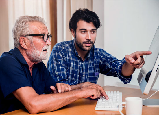  young man in a blue checkered shirt is sitting at a desk next to an older man with glasses and a navy blue shirt.