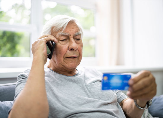 An elderly man with white hair wearing a light gray shirt is sitting on a couch, holding a credit card in one hand and speaking on a mobile phone with the other. 