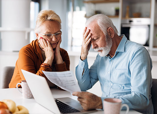 An elderly couple sits at a table with a laptop, looking worried. The man holds a piece of paper and rests his hand on his forehead