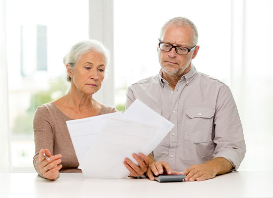 A senior woman with white hair and a tan sweater holds several documents, looking serious. Beside her, a senior man with glasses and a beige shirt examines the life insurance papers, one hand on a calculator. They are seated at a white table in a bright room.
