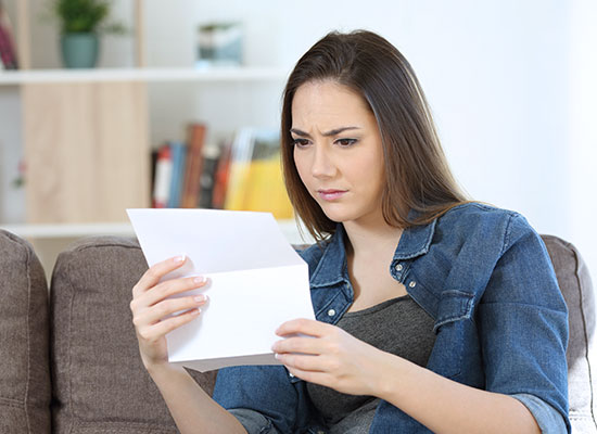 A woman with long brown hair, wearing a denim shirt, sits on a couch. She holds a piece of paper with a concerned expression, aware of the potential inheritance scam it might unveil. A bookshelf is blurred in the background.