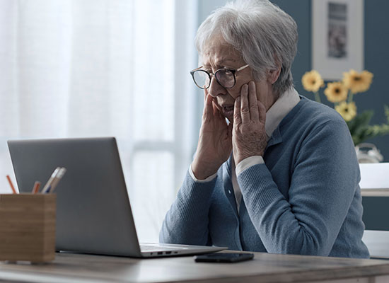 An elderly person with glasses sits at a table, looking surprised at a laptop screen, possibly reading about an inheritance scam. 