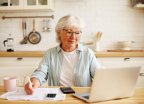 An older woman works at her computer in her kitchen, illustrating aging in place and the importance of community connections and support networks for older Americans living alone.