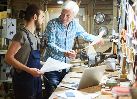 An older man mentoring a younger worker in a workshop, symbolizing intergenerational knowledge transfer in retirement planning.