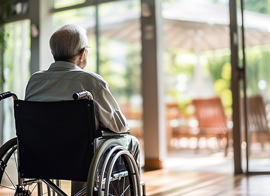 An elderly man in a wheelchair looks out the window, reflecting the challenges of aging solo