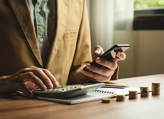 Person in business attire uses calculator and smartphone while working with finances, with neat stacks of coins arranged on a wooden desk.
