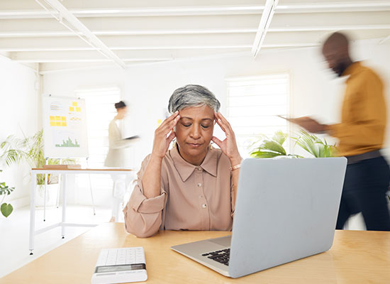 An older woman sits at a desk with her hands on her temples, looking stressed while working on a laptop, representing post-retirement work, retirement challenges, and mental health in retirement.