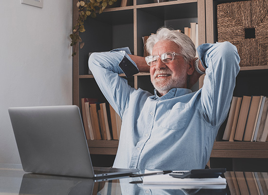 A smiling elderly man relaxing at his desk with a laptop, representing comfortable retirement.