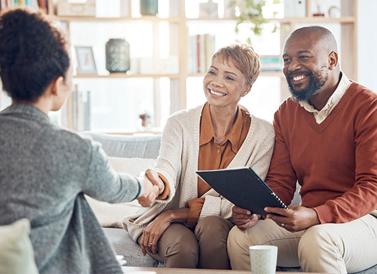A cheerful couple shakes hands with a person across from them in a cozy living room, holding a retirement document and sharing a happy moment.