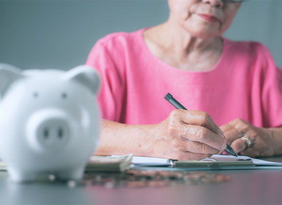 An elderly woman in a pink blouse writes in a notebook, focusing on retirement planning, indicated by a white piggy bank and scattered coins in the foreground.