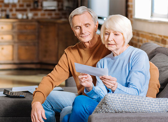 An elderly couple sits on a couch, reviewing a document together. The man, wearing a brown sweater, looks at the paper over the woman's shoulder. The woman, dressed in a light blue sweater, holds the document on social security and taxes. A remote control and other papers are on the couch.