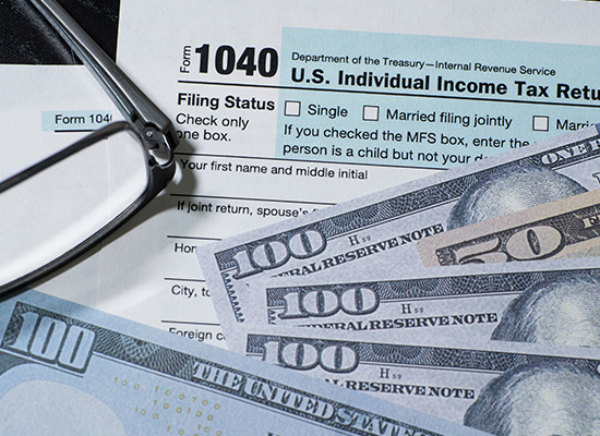 Close-up of a 1040 tax form with glasses and several hundred-dollar bills, representing personal income tax filing.