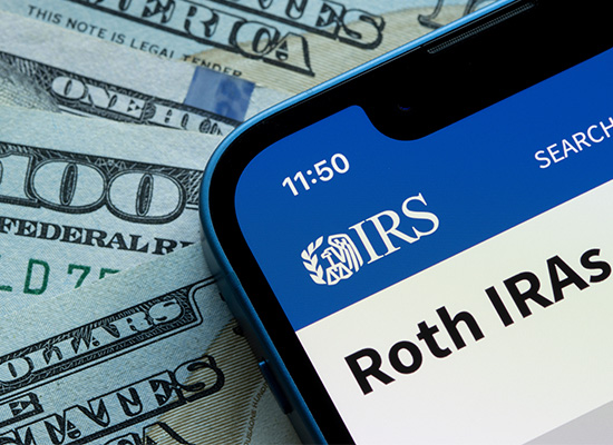 Smartphone displaying the IRS website for Roth IRAs placed over hundred-dollar bills, indicating retirement savings and tax information.