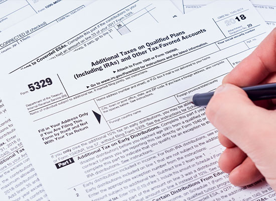 A close-up of a person's hand holding a pen and filling out IRS Form 5329. The form, titled 'Additional Taxes on Qualified Plans (Including IRAs) and Other Tax-Favored Accounts,' is surrounded by other tax documents, worksheets, and information on required minimum distributions.