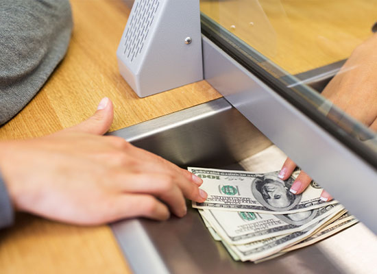 A person's hand passes a stack of U.S. dollar bills through a teller window to another person's hand on the opposite side, who is receiving the money. They are separated by a glass partition in a bank, possibly finalizing details related to required minimum distributions and taxes.