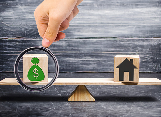 A hand holds a magnifying glass over a scale balancing a money bag and a house