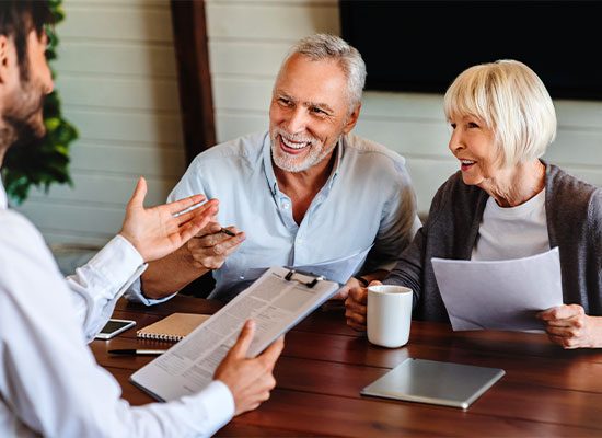 An elderly couple sits at a table discussing retirement planning with a professional holding a clipboard. The man is smiling and gesturing, while the woman holds a document and a mug