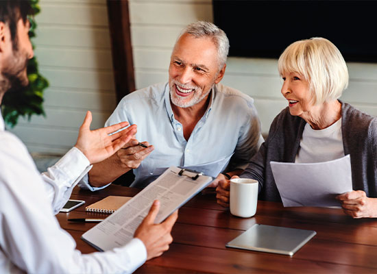 An older couple is joyfully conversing about tax planning with a professional across a table. The man holds a pen, and the woman holds a document. 